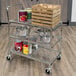 A Luxor metal utility cart with wire shelves holding food containers and a crate on top.