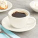 An Acopa Ivory stoneware coffee cup filled with coffee next to a plate of pastries on a table.