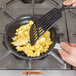 A person using a Vollrath Arkadia aluminum non-stick fry pan to cook scrambled eggs with a spatula.