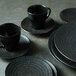 A black Oneida Urban porcelain saucer with a black cup on a table with a stack of black plates.