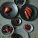 A group of Oneida Urban black porcelain coupe plates with different fruits and berries on them.