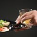 A hand holding a black WNA Comet Milan plastic cocktail plate with a cup holder filled with food and a glass of wine.