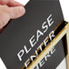 A hand using a Lancaster Table & Seating gold stanchion sign frame to hold a vertical sign that says "Please Enter"