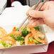 A person holding Town stainless steel chopsticks over a plate of shrimp and broccoli.