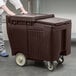A person pushing a Cambro dark brown plastic container with wheels.