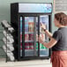 A woman opening an Avantco black countertop display refrigerator with a sliding glass door.