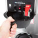 A hand using a Bloomfield automatic coffee maker to open a black container with a red handle.