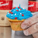 A hand holding a Hoffmaster white fluted baking cup with a cupcake with blue frosting and red and white stars.