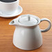 An Arcoroc white teapot on a table with a cup of coffee.