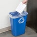 A hand putting paper into a blue Rubbermaid recycling bin.