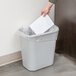 A hand putting a piece of white paper into a Rubbermaid gray rectangular trash can.