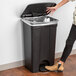A woman opening the black rectangular lid of a Lavex step-on trash can.