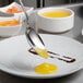 A Mercer Culinary stainless steel plating spoon over a bowl of yellow liquid.