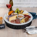 A Frilich black wicker cooling plate with cheese, meats, and wine on a table.