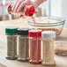 A hand pouring seasoning into a 53/485 round plastic spice container with a red and white dual flapper lid.