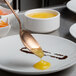 A Mercer Culinary rose gold plating spoon with a bowl of yellow liquid being poured onto a white plate.