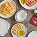 A table with plates and bowls of food including a bowl of pasta with shrimp on a table set with stacks of white Acopa oval bowls.