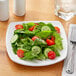 A plate of salad with tomatoes, cucumbers, and lettuce on a white Acopa square porcelain plate.