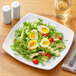 A white Acopa square porcelain plate with a salad, hard boiled eggs, and vegetables.