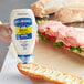 A hand holding a white Hellmann's mayonnaise bottle over a sandwich.