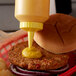 A burger with mustard being poured from a yellow Vollrath squeeze bottle.