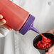 A person using a Vollrath Traex wide mouth squeeze bottle with purple cap to pour sauce into a cup of food.