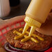 A burger with mustard on a bun held by a Vollrath Twin Tip squeeze bottle with a yellow cap.