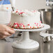 A person using an Ateco round cake turntable to decorate a white cake with red and pink hearts.