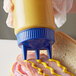 A hand using a Vollrath Tri Tip squeeze bottle with a blue lid to pour mustard onto a sandwich.