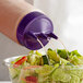 A clear Vollrath Tri Tip wide mouth squeeze bottle with a purple cap pouring dressing onto a salad.