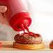 A hand using a Vollrath Twin Tip Squeeze Bottle with red cap to pour ketchup on a hamburger.