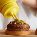 A person pouring mustard onto a burger using a yellow Twin Tip wide mouth bottle cap.