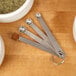 A Tablecraft stainless steel spice measuring spoon set on a wooden table with spices in them.