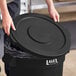 A person places a Lavex black round commercial trash can lid on a black trash can.