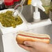 A person holding a hot dog and using a Carlisle condiment dispenser for relish.