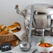 A silver pot with a American Metalcraft hammered stainless steel ladle next to a bowl of bread.