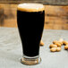 A Libbey pub glass filled with dark beer on a table with peanuts.