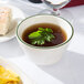 A Tuxton green speckle china bouillon cup filled with brown liquid and a leafy green garnish on a table with a plate of food.