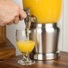 A hand pouring Tablecraft 1.25 gallon stainless steel beverage dispenser with infuser and ice core into a glass of orange juice.