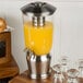 A Tablecraft stainless steel and plastic beverage dispenser with orange juice and glasses.