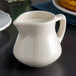 A Tuxton eggshell white china creamer with a handle sits on a table.