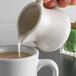 A person pouring milk into a cup of coffee using a Tuxton eggshell china creamer.