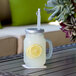 A clear plastic drinking jar filled with lemonade and ice with a lemon slice and a straw on a table.