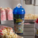 A blue "Fun at the Fair" Tizzeroo souvenir cup with a blue lid and straw next to a bag of popcorn.
