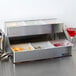 A San Jamar stainless steel condiment bar with food in it next to a glass of red liquid.