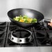 A hand using a Winco wok ring to cook vegetables in a wok on a stove.