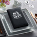 A black H. Risch, Inc. Oakmont menu on a plate on a table with silverware and a white salt shaker.