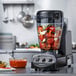 A Vitamix blender with vegetables in a glass container with red sauce.