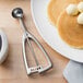 A white plate with a black edge holding pancakes with butter balls and a Vollrath stainless steel squeeze handle disher.