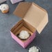 A Baker's Mark pink cupcake box with a cupcake inside and white frosting on top.